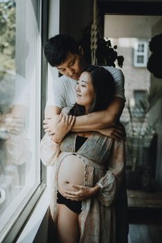 a man and woman embracing each other in front of a window with their arms around the pregnant belly