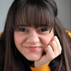 a girl with long brown hair and bangs smiles at the camera while wearing a yellow jacket