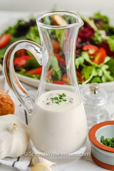 a glass pitcher filled with dressing next to a bowl of vegetables and bread on a table