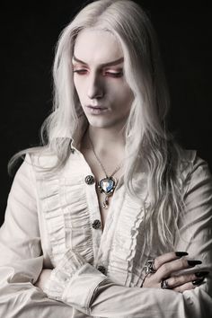 a woman with long white hair wearing a necklace and holding her arms crossed in front of her chest