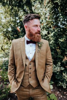 a man with a beard wearing a tan suit and bow tie standing in front of trees