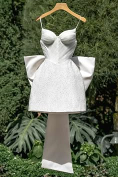 a white dress hanging on a wooden hanger in front of some bushes and trees