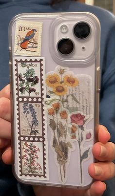 a person holding up a phone case with stamps on it and flowers in the back