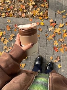 a person is holding a coffee cup in their hand while sitting on the ground with autumn leaves around them