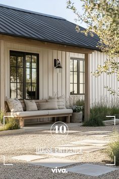 a white house with a black roof and some plants on the front porch, next to a gravel path