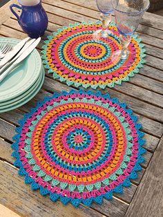 two crocheted placemats sitting on top of a wooden table next to plates