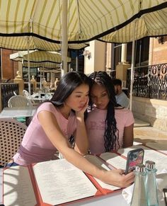 two young women sitting at an outdoor table with open books on it, looking at a cell phone