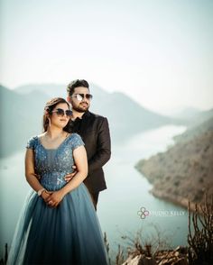 a man and woman standing next to each other on top of a hill near water