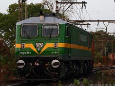 a green and yellow train traveling down tracks next to some power lines with trees in the background