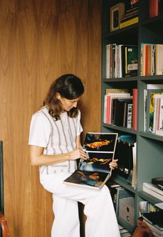 a woman sitting in front of a bookshelf holding an open book and looking at it