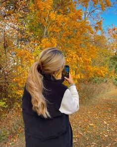 a woman taking a photo with her cell phone in front of some trees that have yellow leaves