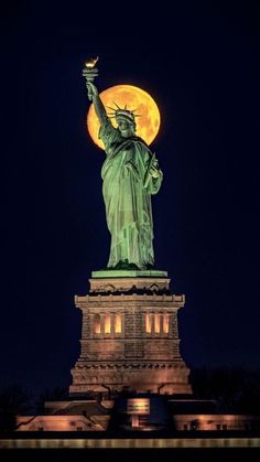 the statue of liberty lit up at night with the full moon in the sky behind it