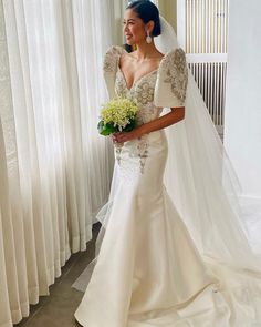 a woman standing in front of a window wearing a wedding dress and holding a bouquet