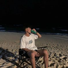 a man sitting in a folding chair drinking from a bottle on the beach at night