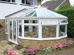 a white sun room sitting in the middle of a garden next to a house with pink flowers