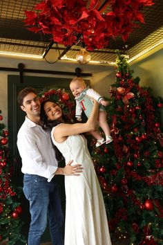 a man and woman holding a baby in front of a christmas tree with poinsettis