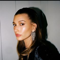 a woman with long hair wearing earrings and posing for the camera in front of a white wall