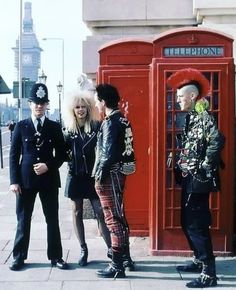 three people standing in front of a red phone booth on the sidewalk with one person dressed as punk