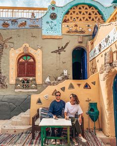 a man and woman sitting on a bench in front of a building with colorfully painted walls