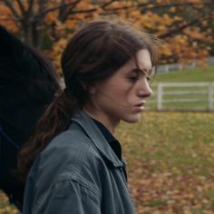 a woman with long hair walking in front of a fence and tree filled field holding a black horse