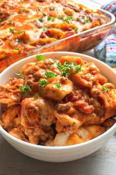 a white bowl filled with pasta and meat covered in sauce next to a casserole dish