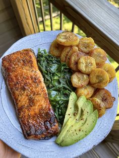 a white plate topped with meat and vegetables next to sliced avocado on a wooden deck