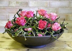 a bowl filled with pink roses sitting on top of a wooden table next to a brick wall