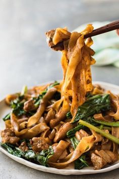 someone is holding chopsticks over noodles with meat and vegetables on a white plate