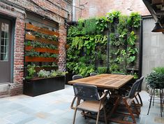 an outdoor dining area with wooden table, chairs and green plants on the wall behind it