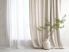 a white vase with a plant in it sitting next to a drapesed window