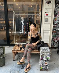 a woman sitting on a wooden bench in front of a store with lots of tattoos