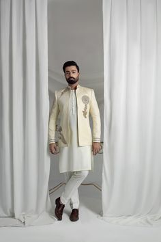 Expertly crafted from jamawar material, this White and Gold Short Prince Coat is adorned with intricate ethnic Sindhi and Rajasthani motifs. The zari, dabka, and crystal embellishments add a touch of opulence. Complete with a silk inner suit, this coat exudes cultural richness and luxuriousness. 3-Piece Suit Festive Embroidered Kundan Sherwani, Gold Traditional Wear With Naqshi Drape, Kundan Sherwani For Eid Festivities, Ceremonial Bandhgala With Zari Work In Raw Silk, Elegant Embroidered Raw Silk Bandhgala, Traditional Gold Raw Silk Bandhgala, Traditional Gold Bandhgala In Raw Silk, Traditional Festive Churidar With Naqshi, Ceremonial Bandhgala With Naqshi Traditional Drape