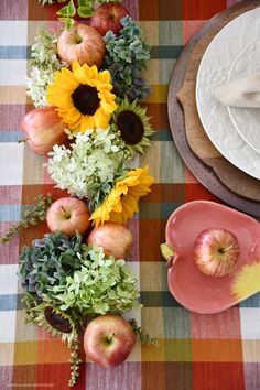 an arrangement of flowers, apples and other fruit on a table with place settings in the background