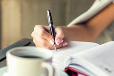 a woman's hand holding a pen and writing on a piece of paper next to a cup of coffee