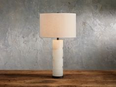 a lamp on a wooden table with a gray wall behind it and a white shade