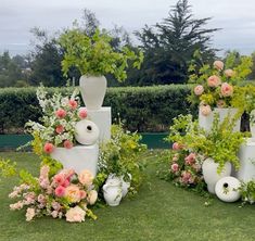 several white vases with flowers and greenery in them on the grass near bushes