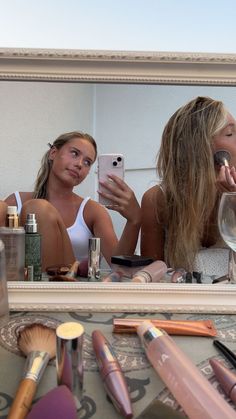 two women looking at their cell phones in front of a mirror with makeup on it