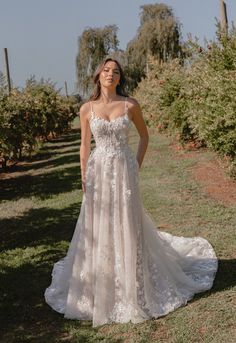 a woman standing in an apple orchard wearing a wedding dress with spaghetti straps and floral appliques