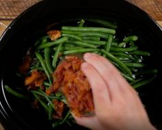 a hand is picking up green beans from a black bowl on a wooden table top