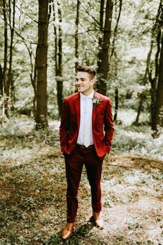 a man in a red suit and white shirt standing in the woods with his hands in his pockets