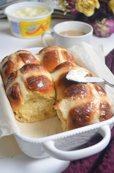 hot cross buns in a white baking dish with butter on top and coffee mugs next to it