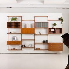 a book shelf with books, plants and pictures on it in an empty room that appears to be white