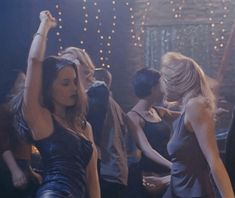 two women dancing at a party with other people in the back ground and lights behind them