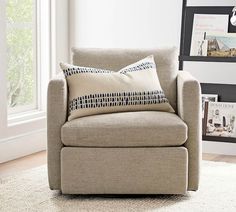 a chair with two pillows on it in front of a window and framed pictures behind it