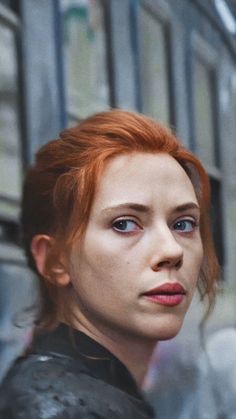 a woman with red hair and blue eyes looking at the camera while standing in front of a building