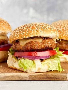 three hamburgers with lettuce, tomato and onion on a wooden cutting board