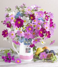 a vase filled with purple and white flowers next to fruit on a wooden table top