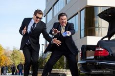 two men dressed in suits and ties jumping into the air next to a black car