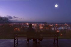 two people are sitting on a bench looking at the city lights in the night sky