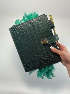 a hand holding an alligator skin notebook with green feathers on the cover and a pen sticking out of it